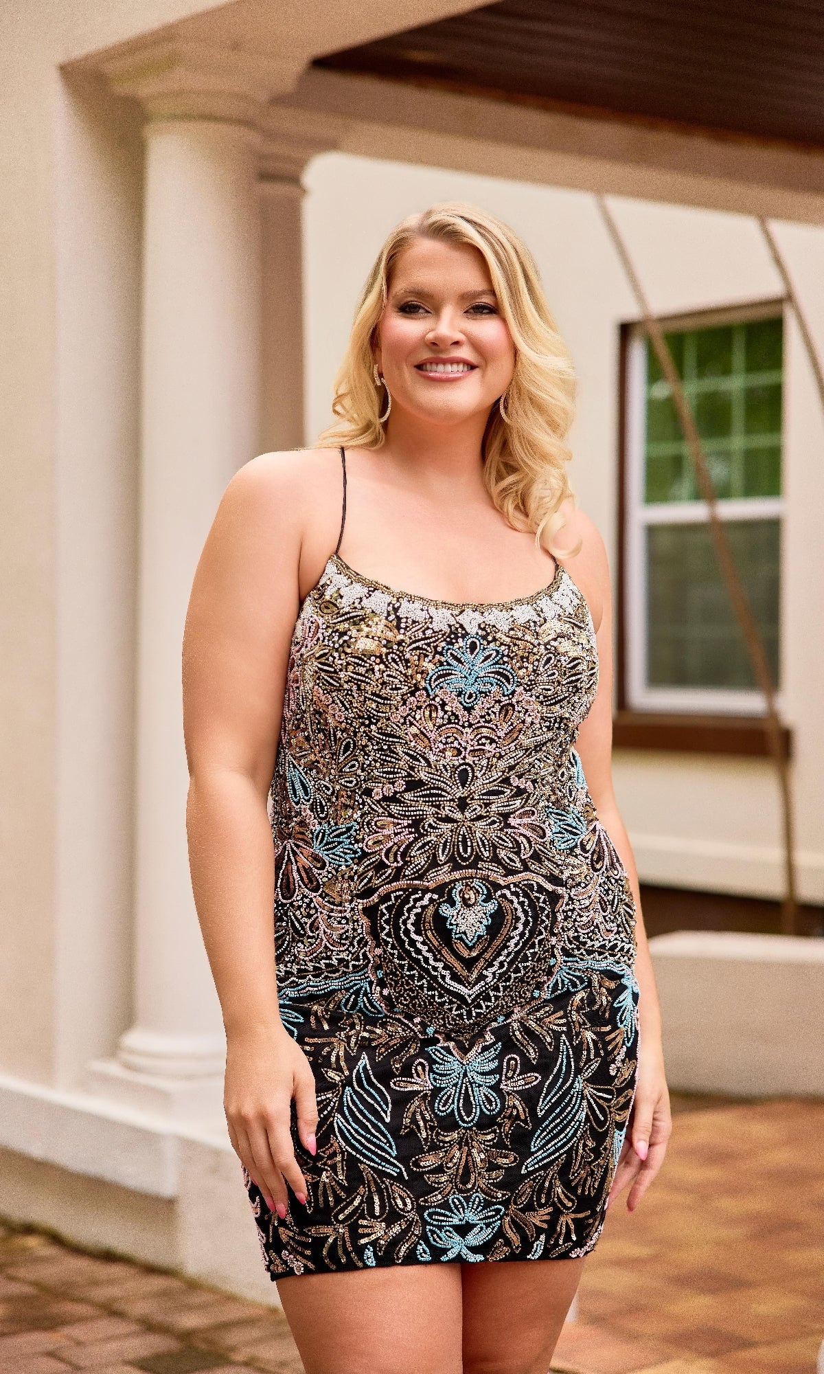 A smiling woman with blonde hair poses outside by a white column and window, wearing the Plus-Size Short Lace-Up Hoco Dress: Primavera 14090.